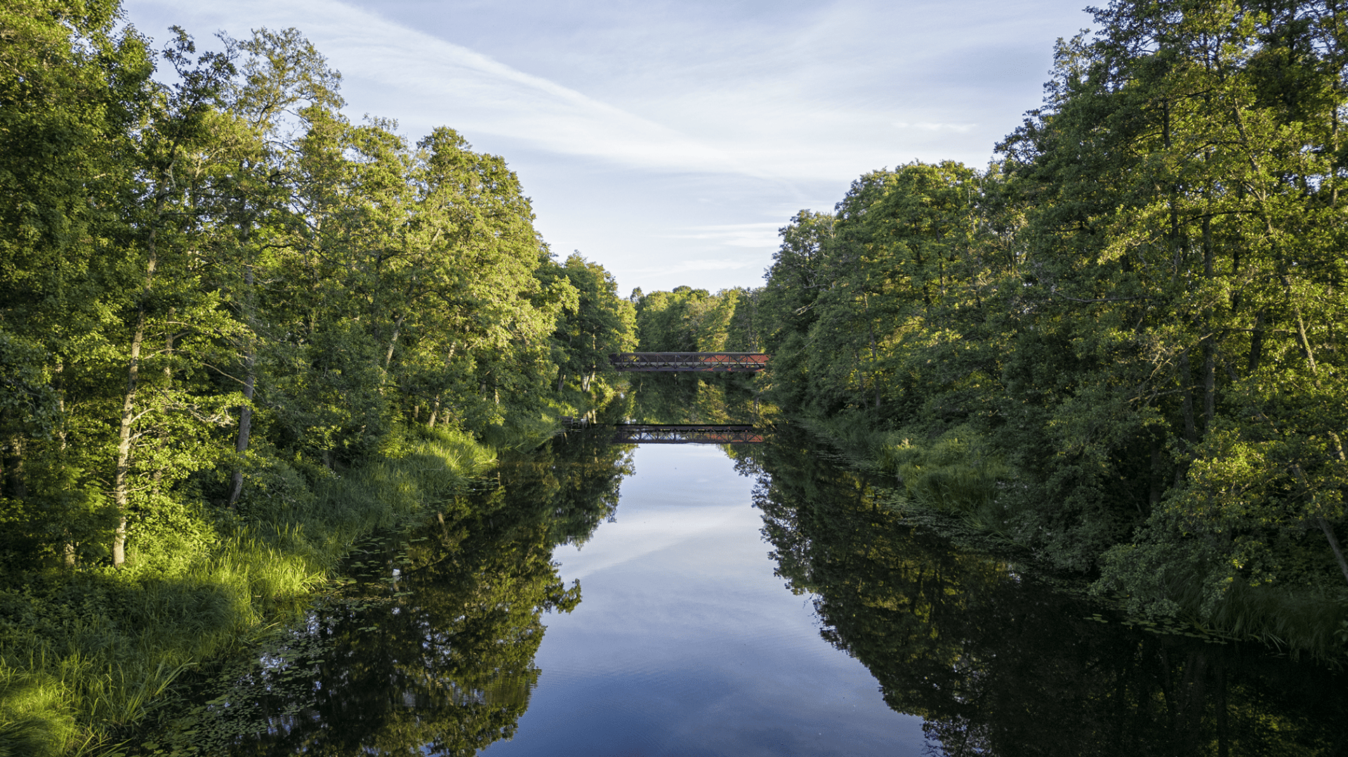 En stilla å med spegelblankt vatten, omgiven av grönskande träd och två träbroar i bakgrunden._